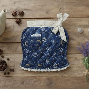 Puede incluir: Una funda para tetera de tela acolchada en color azul marino profundo con un estampado de pequeñas flores blancas y lunas crecientes doradas. El artículo tiene una banda superior color crema con un lazo, un bolsillo pequeño y encaje blanco.