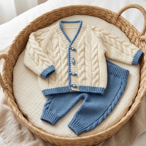 May include: A two-piece knit set consisting of a cream-colored cardigan with cable knit patterns, blue trim, and toggle buttons, paired with matching blue knit pants featuring a vertical cable knit detail along the outer legs.