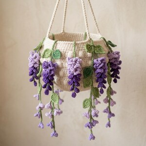 May include: A cream-colored crocheted hanging basket featuring cascading wisteria-like floral vines in shades of light and dark purple. The basket has a textured, rounded body and is suspended by three braided cream-colored yarn cords.
