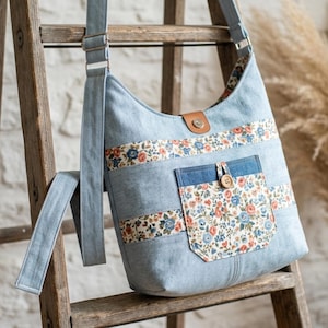 Pode incluir: Uma bolsa hobo de ganga azul-clara com faixas horizontais de tecido floral em tons de laranja, azul e creme. A bolsa inclui um bolso frontal com estampado floral, uma aba de fecho em pele castanha com mola metálica e uma alça ajustável.
