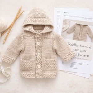 May include: A beige knit hooded cardigan with four buttons and two front pockets, accompanied by two balls of yarn, wooden knitting needles, a measuring tape, and a printed document titled Toddler Hooded Cardigan Knitting Pattern.