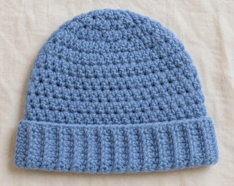 May include: A light blue beanie hat crafted with a textured crochet stitch pattern. The hat features a wide, ribbed brim at the base and a rounded crown, showcasing a consistent yarn color and uniform hand-stitched construction throughout.