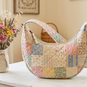 Puede incluir: Un bolso hobo en forma de media luna hecho de tela acolchada con un diseño de patchwork de varios patrones florales y de rayas en tonos pastel suaves de rosa, azul, amarillo y crema, con una correa a juego y cierre de cremallera superior.