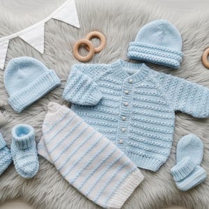 May include: A light blue knitted baby set including a long-sleeve button-up cardigan, two beanies, two pairs of booties, and a pair of mittens. The set is accompanied by a white and blue striped blanket, a small grey plush elephant, and wooden rings.