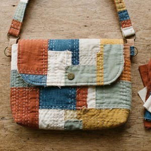 Peut inclure: Un sac à bandoulière patchwork rectangulaire doté d'une texture matelassée avec des points de piqûre blancs visibles. Le sac est composé de carrés de tissu dans des tons orange rouille, bleu marine, crème, vert sauge et jaune moutarde, fermé par un bouton-pression en laiton.