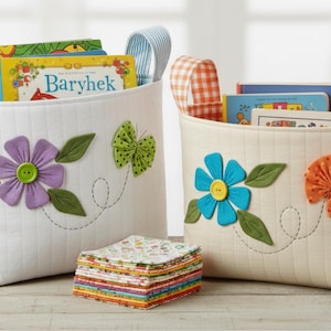 Peut inclure: Deux paniers de rangement en tissu avec des appliqués floraux et papillons. L'un est blanc avec une fleur violette et un papillon vert, l'autre est crème avec une fleur bleue et un papillon orange. Les deux sont remplis de livres.