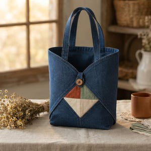 May include: A dark blue denim tote bag featuring two sturdy handles and a central button closure. The front panel displays a diamond-shaped patchwork design composed of four triangular sections in rust, sage green, and cream colors.