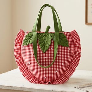 May include: A strawberry-shaped tote bag featuring a red and white gingham pattern, green fabric leaves at the top, and a ruffled border. The bag includes two green handles and small white embroidered dots scattered across the red gingham surface.