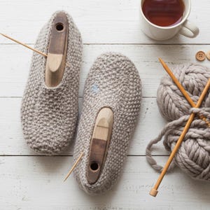 May include: A pair of beige knitted slippers featuring wooden shoe trees inside, accompanied by a ball of thick beige yarn, two wooden knitting needles, and several small wooden buttons arranged on a white wooden surface.