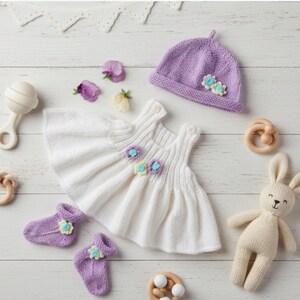May include: A white knitted dress with floral accents, a purple knitted hat with flowers, two purple knitted booties with floral details, a beige knitted rabbit toy, a cream rattle, and two wooden teething rings arranged on a white surface.