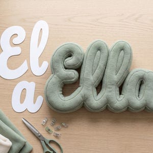 May include: A sage green fabric letter pillow spelling the name Ella in cursive script, accompanied by white paper cutouts of the letters E, l, a, a, a spool of green thread, metal sewing clips, scissors, and folded fabric pieces.