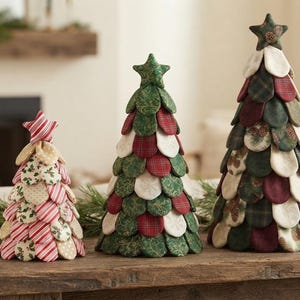 Puede incluir: Tres árboles de Navidad decorativos de tela de diferentes alturas, construidos con piezas de tela superpuestas en forma de pétalo con patrones rojos, verdes, crema y de cuadros, cada uno rematado con una pequeña estrella de tela.