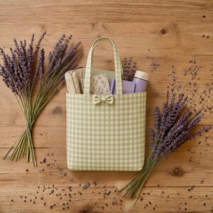 Puede incluir: Un bolso tote de cuadros vichy en verde claro y blanco con un pequeño lazo a juego en la parte frontal. El bolso contiene un cuaderno, un estuche pequeño y una botella de color violeta claro. Dos ramos de lavanda seca violeta flanquean el bolso.