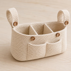 May include: A beige crochet storage basket featuring two hanging loops, two exterior front pockets, and a central divider. The basket is accented with four small, round, brown wooden buttons attached to the loops and the front panel.