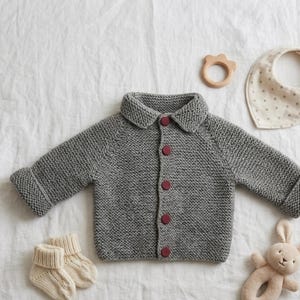 May include: A gray knitted baby cardigan with burgundy buttons, a matching pair of socks, a wooden teether, a dotted bib, and a stuffed bunny toy are arranged on a white surface. The cardigan has a classic collar and rolled cuffs.
