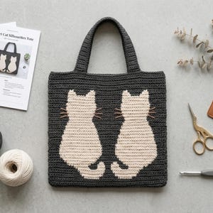 May include: A dark gray crochet tote bag featuring two cream-colored cat silhouettes on the front. The bag has two handles and is displayed alongside a printed pattern titled Crochet Cat Silhouettes Tote, yarn balls, scissors, and a crochet hook.