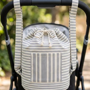 May include: A drawstring backpack featuring a grey and white horizontal striped pattern. The bag includes two matching striped shoulder straps, a front pocket with vertical stripes, and a white cord closure with knotted ends at the top opening.