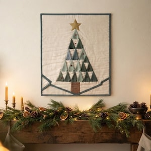 Könnte beinhalten: Ein quadratischer, gesteppter Wandbehang mit einem stilisierten Weihnachtsbaum aus dreieckigen Stoffstücken in Grün- und Blautönen. Der Baum ist mit einem goldenen Stern gekrönt und hat einen braunen Stamm auf cremefarbenem Grund.