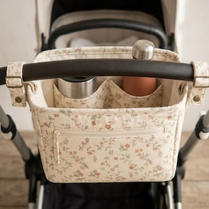 Puede incluir: Un organizador de cochecito rectangular de tela color crema con un pequeño estampado floral en tonos rojos y verdes apagados. Incluye un bolsillo frontal con cremallera y dos compartimentos internos que contienen una botella de metal y una botella color melocotón.