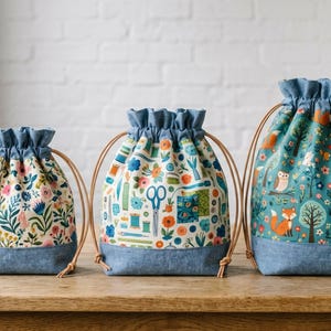 May include: Three drawstring bags in varying sizes with blue denim-style bases and ruffled tops. The smallest features a floral print, the middle shows sewing tools and flowers, and the largest displays a forest scene with foxes and owls.