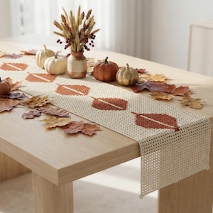 May include: A cream-colored crocheted table runner featuring a repeating pattern of rust-orange leaf shapes. The runner is decorated with small white and orange pumpkins and scattered dried autumn leaves, with a vase of dried wheat stalks.