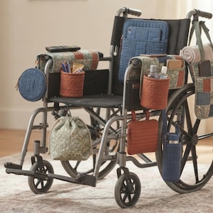 Peut inclure: Un fauteuil roulant gris équipé de divers accessoires de rangement en tissu matelassé, notamment des organisateurs d'accoudoirs, une pochette de dossier, un sac latéral suspendu, une bourse, un porte-bouteille et une petite poche carrée.
