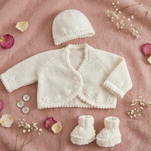 May include: A white knitted baby set consisting of a long-sleeved cardigan with three buttons, a matching beanie hat, and a pair of booties. The cardigan, hat, and booties feature small, delicate knitted flower details along the edges.