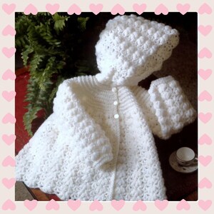 May include: A white, crocheted baby outfit, including a hooded jacket and matching mittens. The jacket has a button closure and a textured pattern. The set is displayed on a dark surface with a fern in the background.