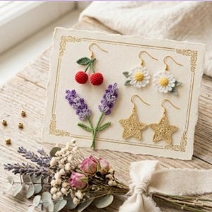 May include: A rectangular display card featuring three pairs of crocheted earrings: red cherries with green leaves, white daisies with yellow centers, and gold stars. The card has a decorative gold border and is presented with a small dried flower bouquet.