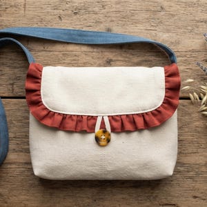 May include: A cream-colored crossbody bag featuring a rust-colored ruffled trim along the flap, a single brown tortoiseshell-patterned button closure with a fabric loop, and a long blue denim shoulder strap attached to the sides of the bag.