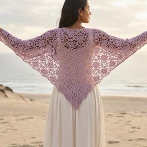 May include: A light lavender crochet shawl featuring an intricate openwork floral pattern with long sleeves. The garment is draped over the shoulders and back, extending outward to form a triangular shape that reaches down to the mid-back area.