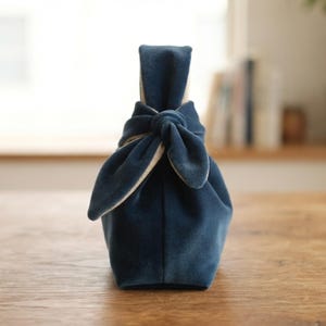 Könnte beinhalten: Eine dunkelblaue Handtasche aus Samt mit einem geknoteten Griffdesign. Die Tasche hat eine weiche, plüschige Textur und einen strukturierten Boden, wobei ein helleres Innenfutter am oberen Knotenverschluss sichtbar ist, was ein schlichtes Accessoire ergibt.