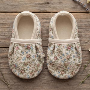 Puede incluir: Un par de zapatos para bebé de suela blanda en tela color crema con un delicado estampado floral de rosas rosadas y pequeñas flores azules. Los zapatos tienen punta redondeada, un panel frontal fruncido y un borde liso de color crema.
