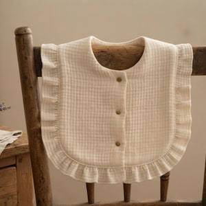 May include: A cream-coloured bib featuring a subtle checkered pattern and a ruffled edge. The bib has a rounded neckline and is secured at the front with three small, metallic-toned snap buttons arranged vertically down the centre.