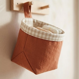Puede incluir: Una cesta de almacenamiento de tela colgante en color naranja quemado sólido con un patrón de cuadros vichy en beige y blanco a lo largo del borde superior. La cesta cuenta con un lazo de tela a juego para colgar y contiene textiles claros.
