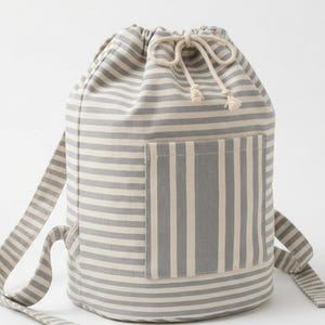 May include: A cylindrical drawstring backpack featuring a grey and white horizontal striped pattern. The bag includes a front pocket with vertical stripes, two matching shoulder straps, and a cream-colored cord closure with knotted ends.