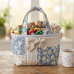 May include: A quilted fabric basket with two handles, featuring a blue and white daisy floral pattern and a solid beige panel with a central fabric bow. The basket is filled with various small wrapped candies and gift boxes.