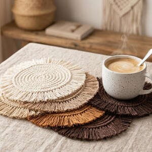 May include: A set of five circular macrame coasters featuring a spiral cord center and a fringed edge. The coasters are arranged in a stack and come in a gradient of colors including cream, beige, tan, mustard yellow, and dark brown.