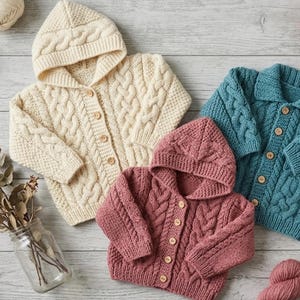 May include: Three knitted cardigans in cream, dusty rose, and teal. The cream and rose cardigans feature hoods and cable knit patterns with wooden buttons. The teal cardigan has a collar, cable knit details, and matching wooden buttons.