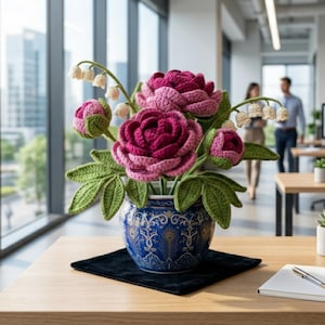 May include: A handcrafted crochet floral arrangement featuring vibrant pink peonies, green leaves, and delicate white lily of the valley. The flowers are displayed in a blue and white patterned vase, resting on a black square, placed on a wooden desk.
