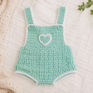 May include: A mint green crochet baby romper with white trim and straps. The romper features a white heart appliqué on the front and two green buttons. The garment is displayed on a white surface.