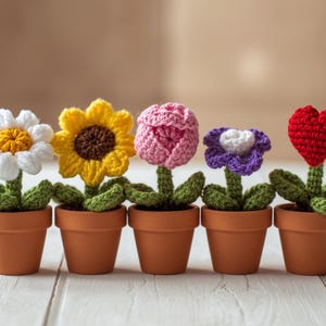 May include: Five crochet flowers in terracotta pots. The flowers include a white daisy, a yellow sunflower, a pink tulip, a purple flower, and a red heart. Each flower has green leaves and stems. The pots are arranged on a white wooden surface.