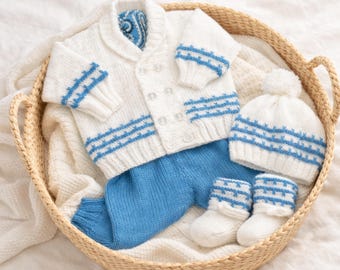 May include: A knitted baby outfit in white and blue, comprising a cardigan, trousers, hat, and booties. The set has blue horizontal stripe detailing on the sleeves, hat, and booties. The outfit is presented in a woven basket.