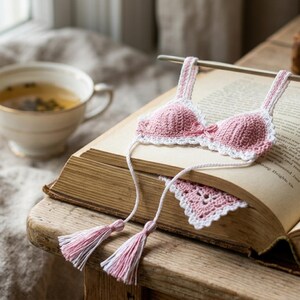 May include: A crochet bookmark shaped like a miniature pink and white bikini top with thin straps and two long white strings ending in pink and white tassels. The piece is resting on the open pages of a vintage book with visible printed text.