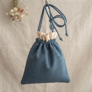 May include: A blue drawstring pouch with a cream-coloured inner lining and a long, matching blue cord strap. The bag features a soft, textured fabric exterior and a gathered top closure that cinches securely with the integrated cord handle.