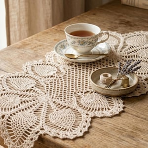 May include: A beige crocheted lace doily with a floral design, a teacup and saucer, a small ceramic bowl with dried lavender, and a ball of yarn on a wooden table. A crochet hook and a spool of thread are also present.