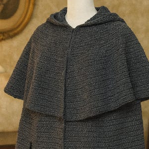 May include: A dark charcoal gray hooded cape featuring a textured, knit-like surface. The garment has a wide, draped collar that extends from the hood, covering the shoulders and upper chest with a consistent, uniform pattern throughout the fabric.