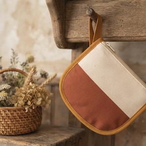 Puede incluir: Un estuche semicircular con un diseño bicolor que presenta una sección superior de color crema y una sección inferior de color terracota. El estuche está ribeteado con un borde amarillo mostaza e incluye una trabilla a juego.