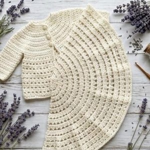May include: A cream-coloured crocheted set consisting of a long-sleeved cardigan with three buttons and a matching circular skirt. The garment features a repeating openwork stitch pattern throughout, showcasing a textured, handmade construction.
