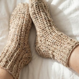 May include: A pair of ankle-length socks crafted from a thick, beige yarn with subtle brown and black flecks. The socks feature a prominent vertical ribbed texture throughout the body, with a reinforced heel and a snug, fitted cuff design.